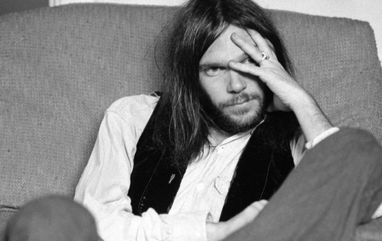 Black and white shot of a young Neil Young with brown shoulder length hair, wearing a white shirt and black waistcoat.