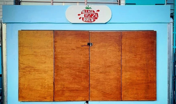 sky blue trailer with a sign that reads "the village pizza" on top. Shop front is boarded up with wood.