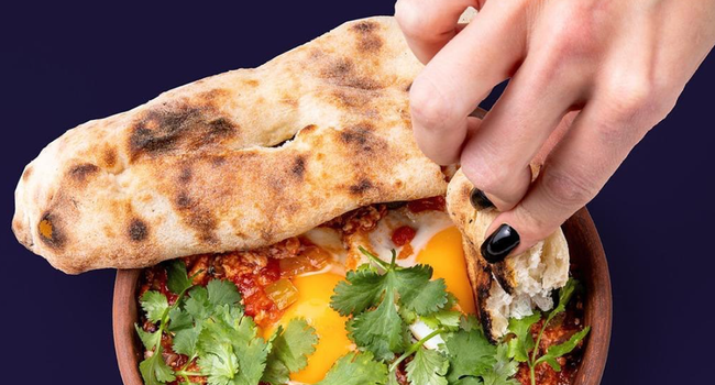 a hand dipping flatbread into a dish with fried egg, herbs and spices.