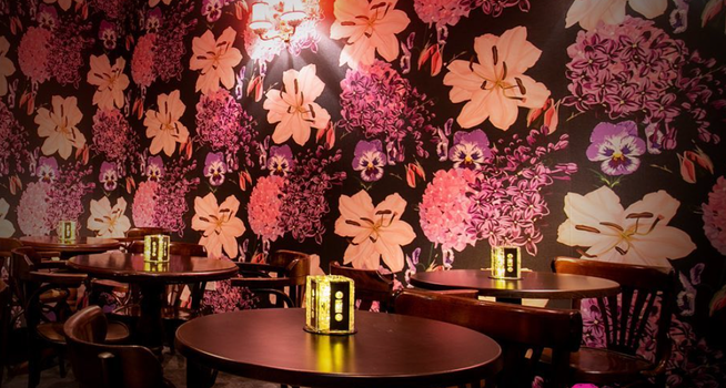 Interior of a bar with pink and brown floral wallpaper and tables and chairs with candleholders made from cassette tapes