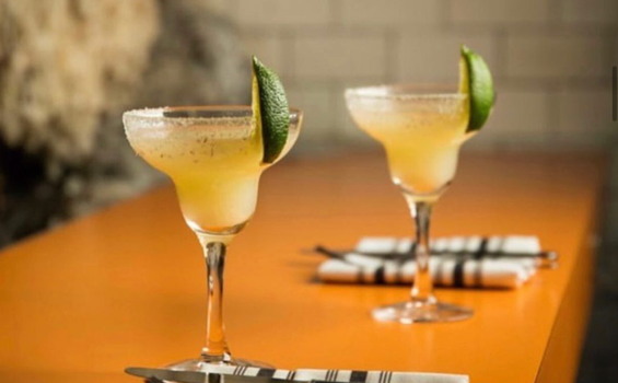 two margaritas with salt rims and fresh lime on an orange table with knives, forks and napkins beside them