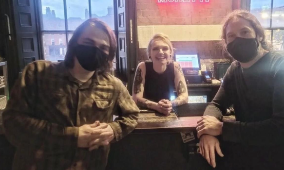 two men in face masks posing in front of a bar, a smiling bartender behind it.