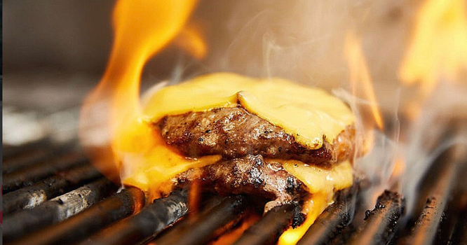 two burger patties on a flaming grill with melted cheese slices on top
