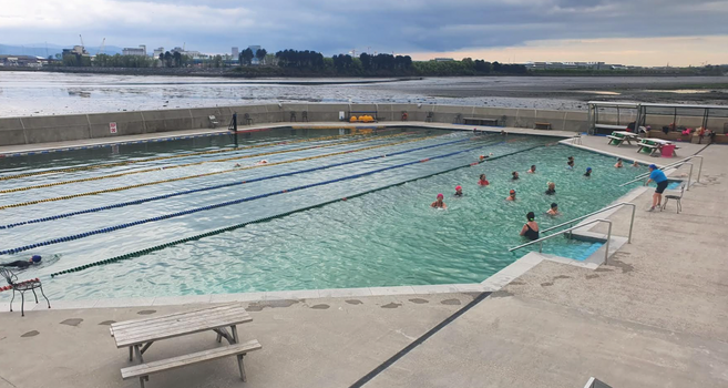 people swimming in an outdoor tidal pool, with views of the irish sea beyond