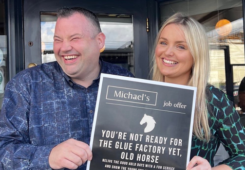 man and woman laughing and holding poster which reads "you're not ready for the glue factory yet, old horse" with Michaels logo above this